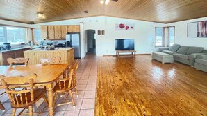 Amplia sala de estar con piso de madera, una mesa de comedor con sillas, cocina con gabinetes de madera, un televisor y un sofá gris.