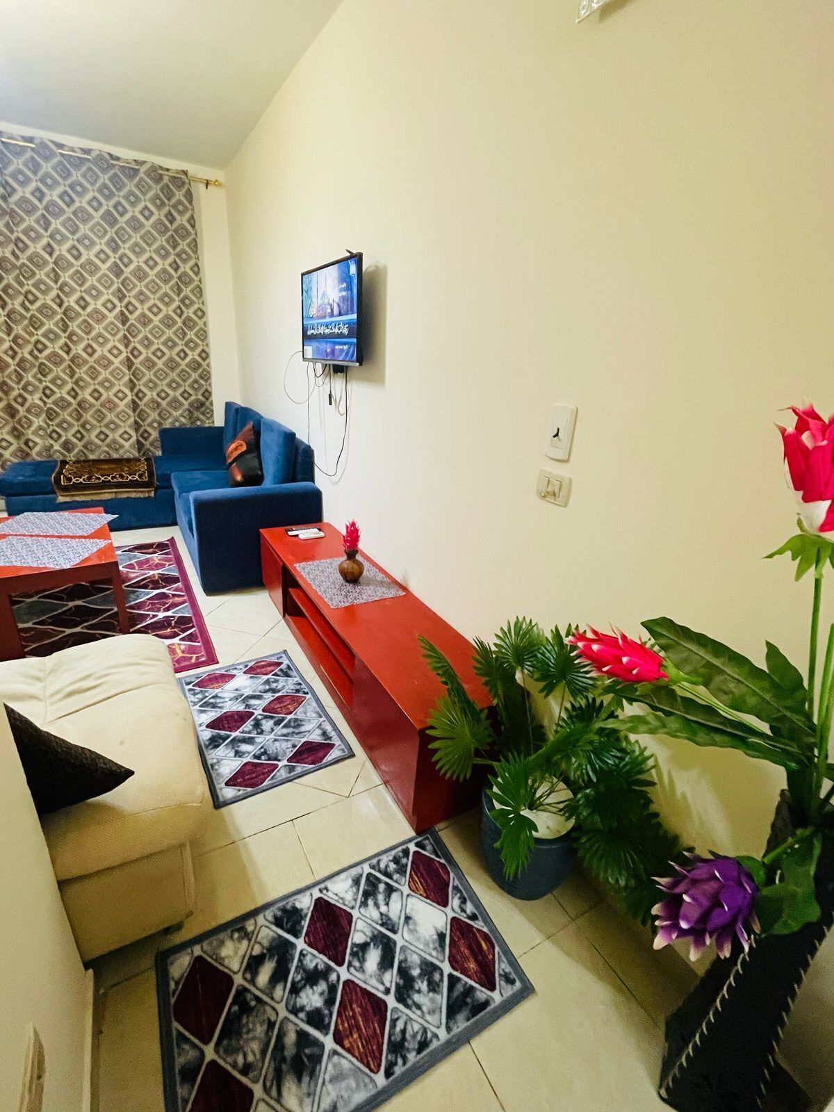A living room with a sofa, a red coffee table, a TV mounted on the wall, and a patterned rug.