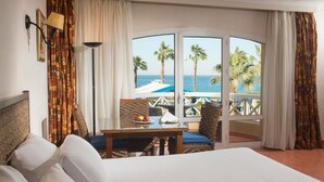 Habitación de hotel con una cama grande, una mesita con fruta, vista al mar y un balcón con piscina.