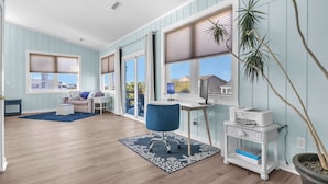 Una habitación con paredes azul claro, suelo de madera, un escritorio con una impresora, una silla de oficina azul, una planta en maceta y una ventana con persianas.