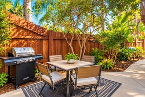 An outdoor patio with a grill, table, chairs, and a potted plant.