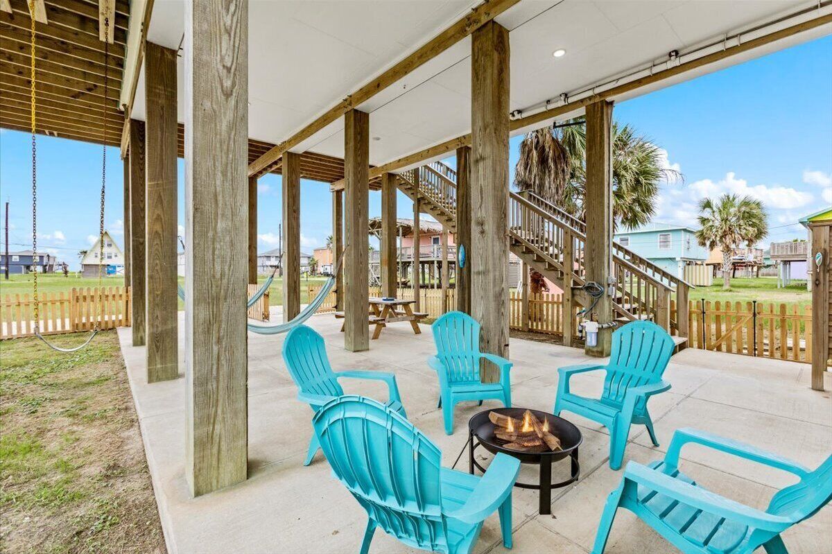 A covered outdoor area with wooden pillars, turquoise Adirondack chairs, a fire pit, and a view of a residential area with houses and a fence.
