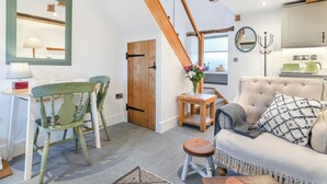 A cozy living room with a high ceiling, a wooden staircase, a dining area with a table and chairs, and a kitchen area with a kettle and a green appliance. - Clarence Cottage - UK47494 (Winkhill, near Leek)