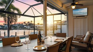 A dining area with a table set for two, overlooking a body of water with palm trees and houses in the background.
