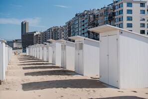 Une rangée de cabanes de plage blanches, avec un chemin de sable menant à elles, se dressant sur fond d’immeubles à plusieurs étages.