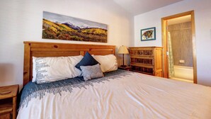 A bedroom with a wooden headboard, a large bed with patterned bedding, a nightstand with a lamp, a wooden dresser, and a painting of a landscape on the wall. - Beautiful Home with views of Lake Dillon, 727B (Frisco)