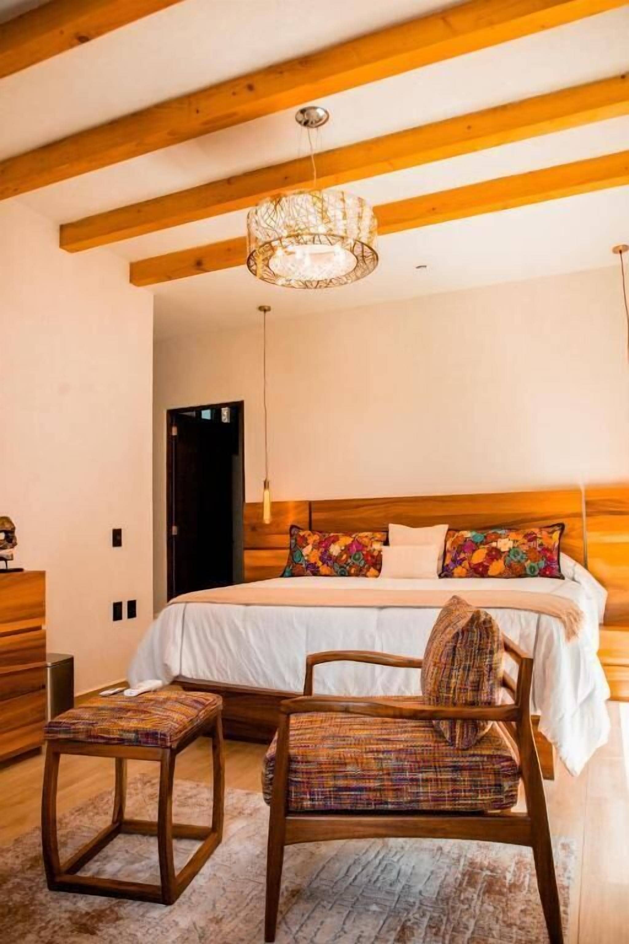 Une chambre à coucher avec une tête de lit en bois, un grand lit agrémenté de coussins colorés, un luminaire suspendu et un banc en bois.
