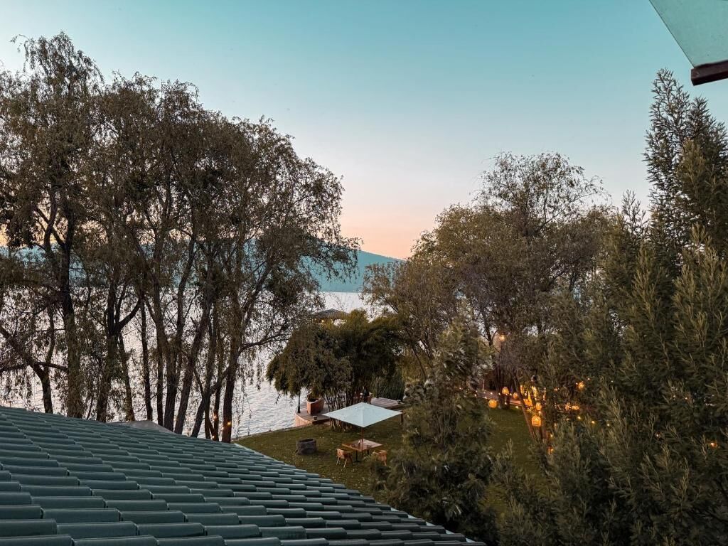 Una terraza con vistas a un lago y árboles, con mobiliario exterior y un edificio al fondo.