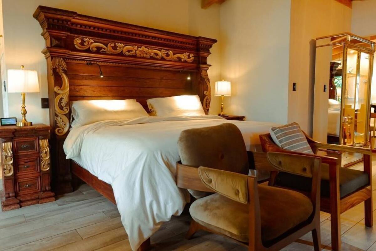 Um quarto espaçoso com uma cama de madeira grande, duas mesas de cabeceira com abajures, uma cadeira de madeira e um espelho.