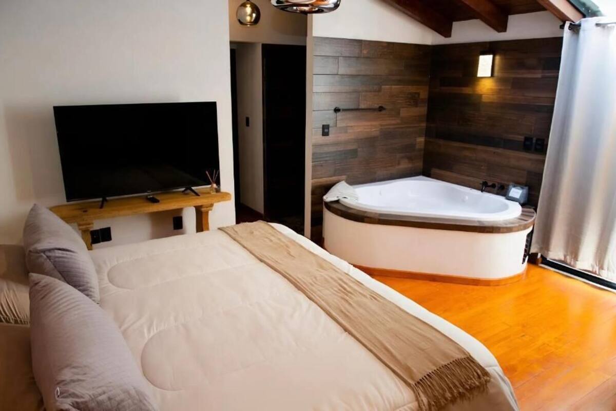 Quarto com cama grande, televisão sobre suporte de madeira e banheira de canto.