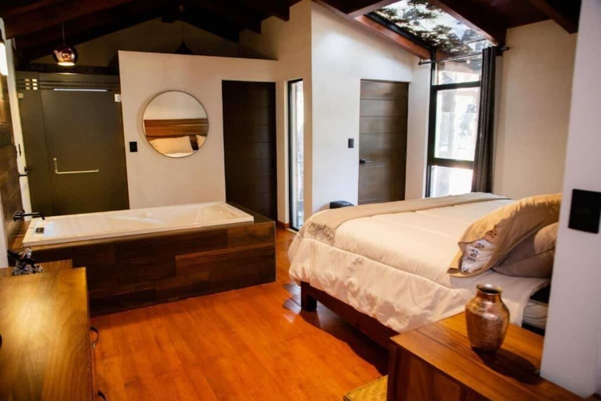 Quarto com cama grande, piso de madeira, espelho circular, banheira integrada e janela com cortinas.