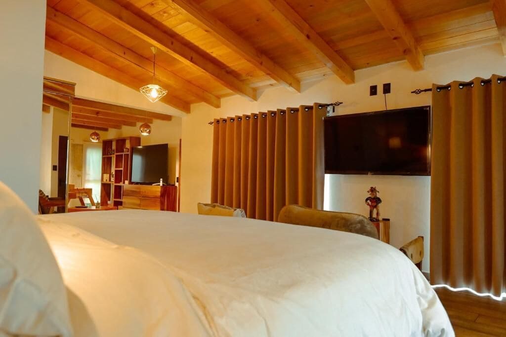 A bedroom with a large bed, wooden ceiling beams, a flat-screen TV, and a window with curtains.