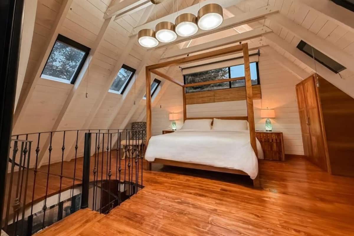 Quarto amplo com teto alto, cama de madeira com dossel, claraboias e escada.