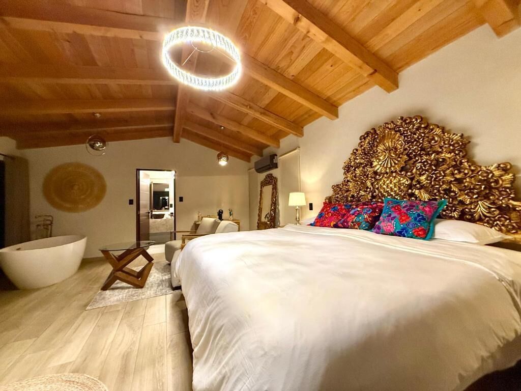 A bedroom with a large bed, a wooden ceiling, a round light fixture, a bathtub, and a mirror.