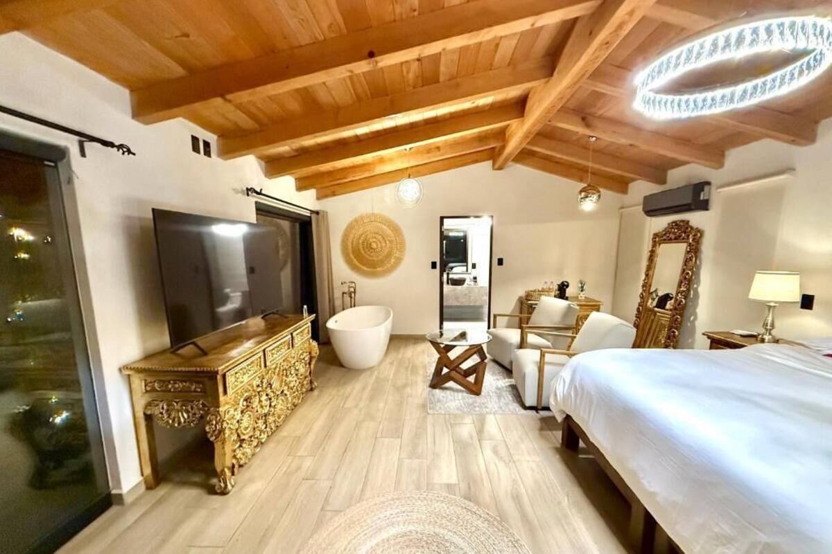 Quarto com teto de madeira, uma cama grande, televisão, banheira branca e área de jantar com cadeiras.