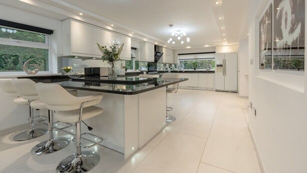 A modern kitchen with white cabinetry, a central island with bar stools, and a large window providing a view of greenery. - Manchester House Retreat (Manchester)