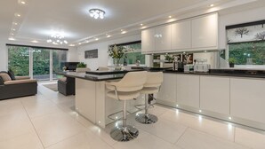 A modern kitchen with white cabinetry, a central island with bar stools, and a large window with a view of greenery. - Manchester House Retreat (Manchester)