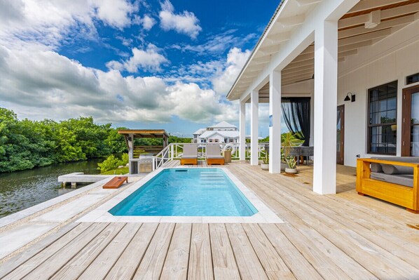 Eine Holzterrasse mit Pool, Liegestühlen und Blick auf ein Haus gegenüber dem Wasser.