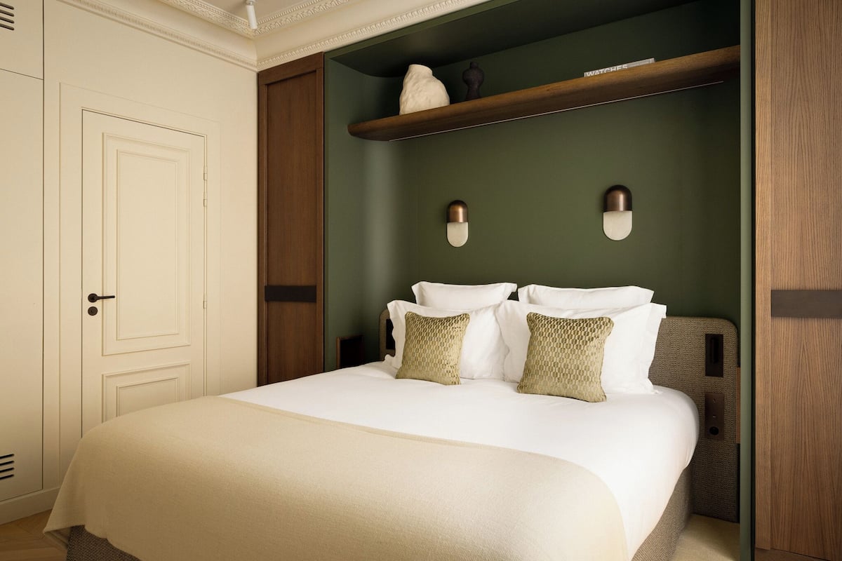 A neatly made bed with white linens and two patterned pillows, a wooden headboard, and a shelf above the bed with decorative items.
