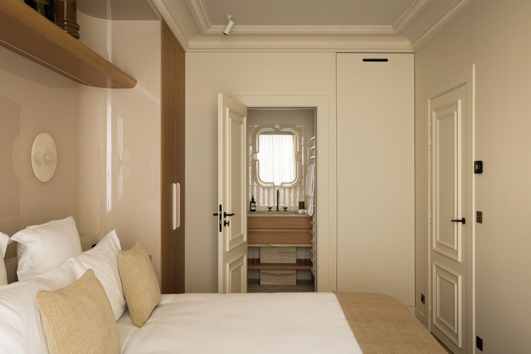 Un dormitorio con cama, almohadas, un estante de madera, una puerta y un baño con ventana y perchero.