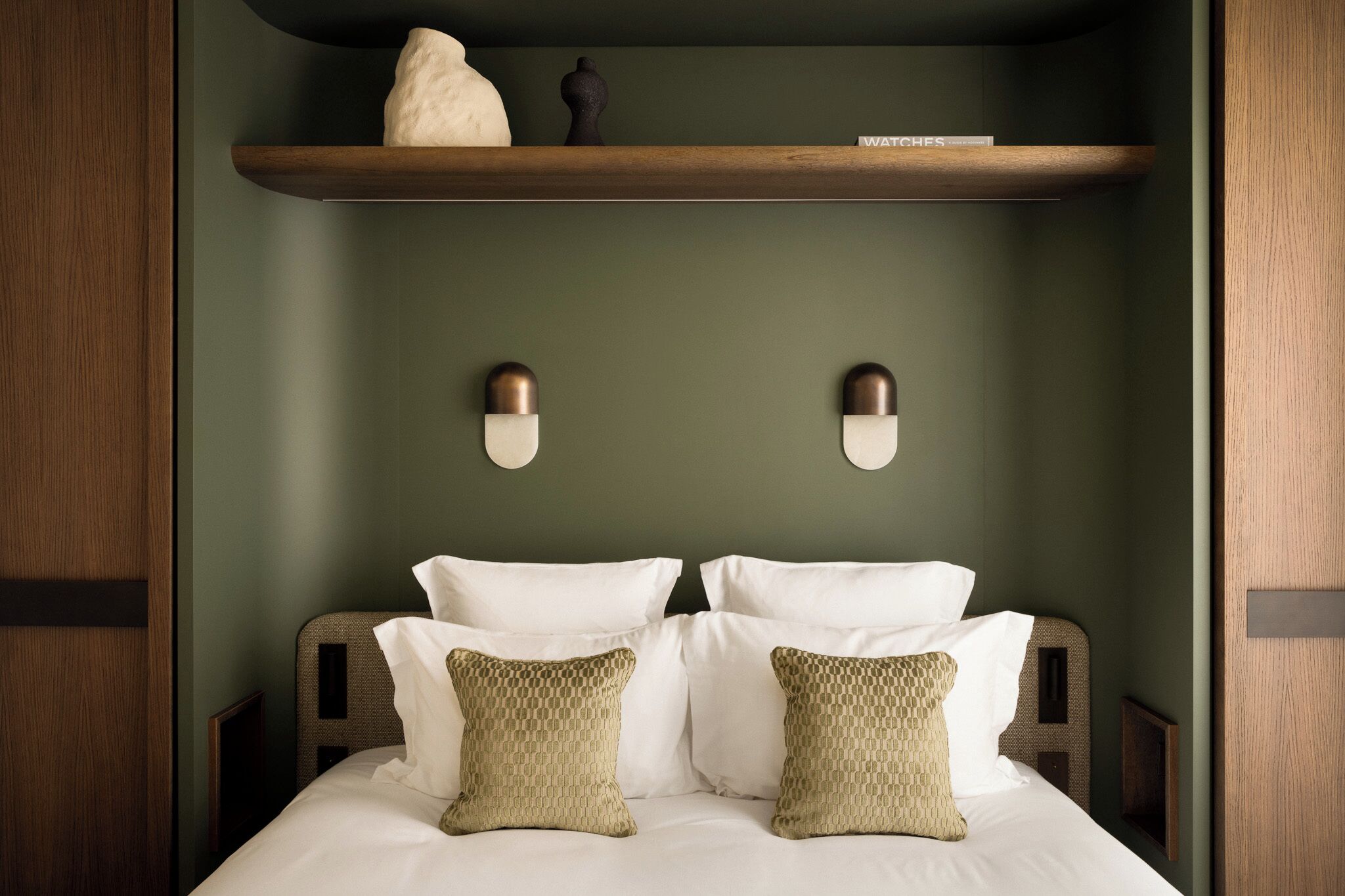Una cama bien hecha con sábanas blancas y almohadas estampadas, un estante de madera arriba y dos lámparas fijadas a la pared.