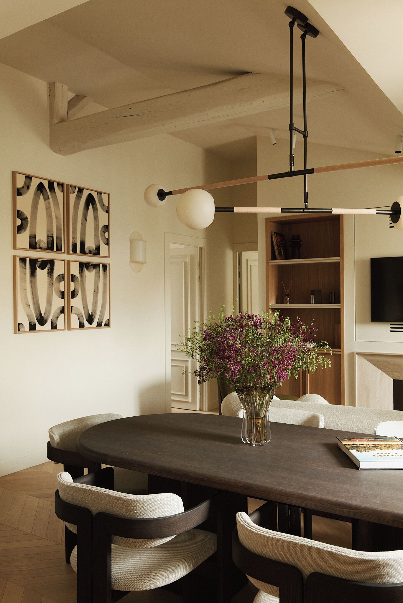A dining area with a dark wooden table, beige upholstered chairs, a vase with flowers, and abstract wall art.
