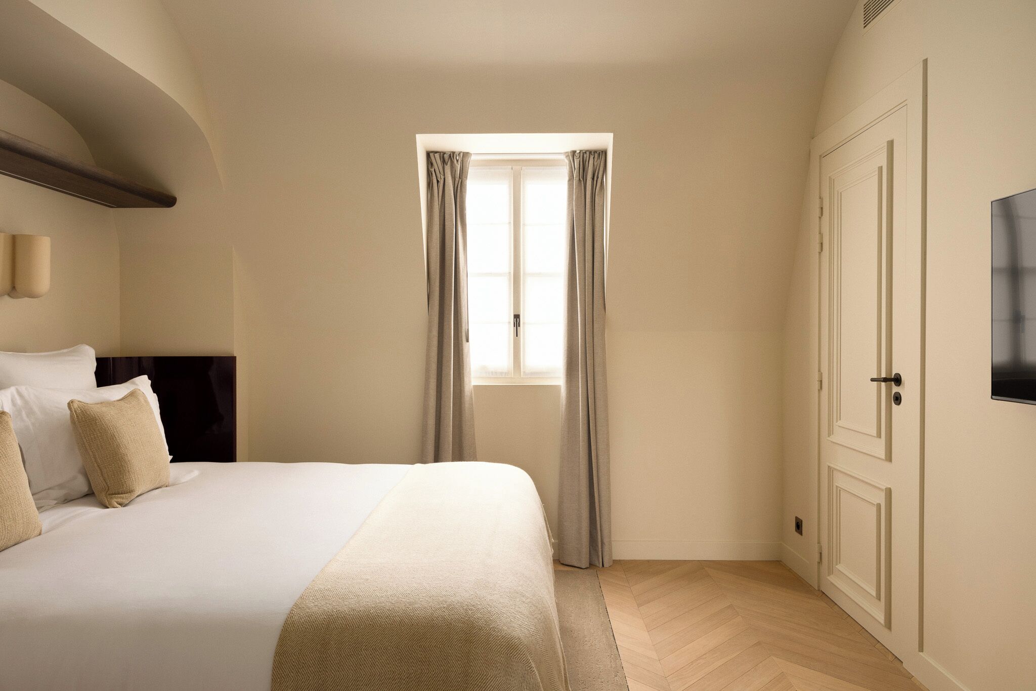 A hotel room with a bed, a window with curtains, a television mounted on the wall, and a door.