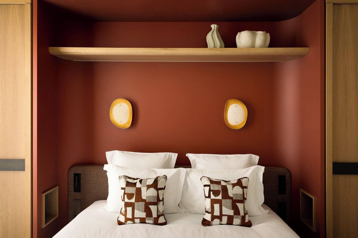A hotel room with a bed, two pillows, a shelf with decorative items, and wall-mounted lights.
