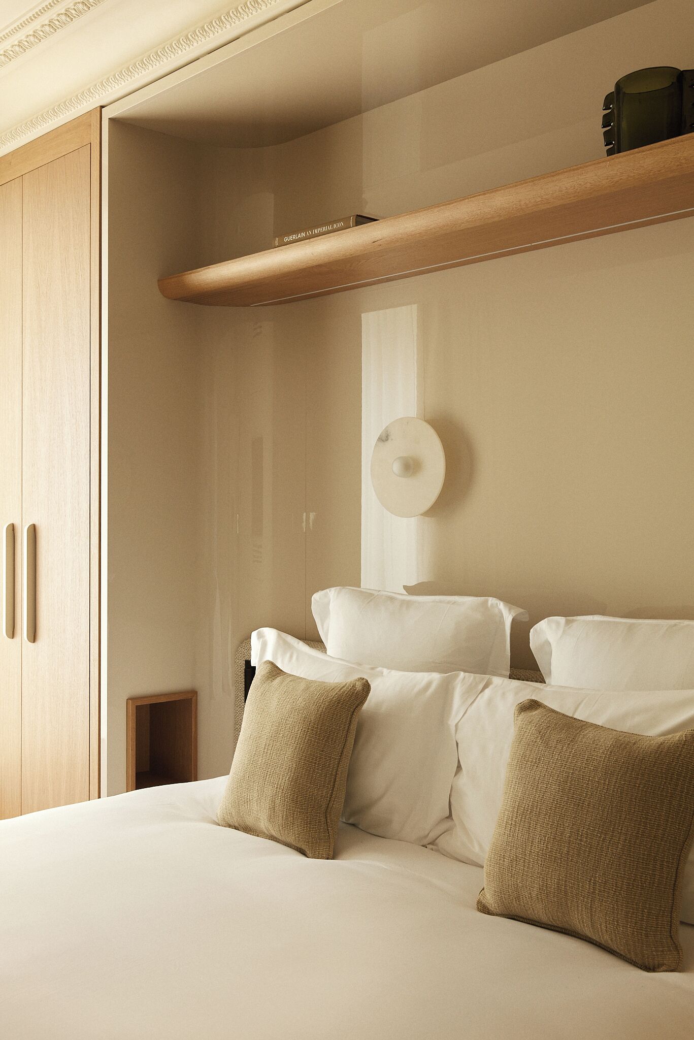 A bedroom with a bed, pillows, a wooden shelf, and a wall-mounted light fixture.