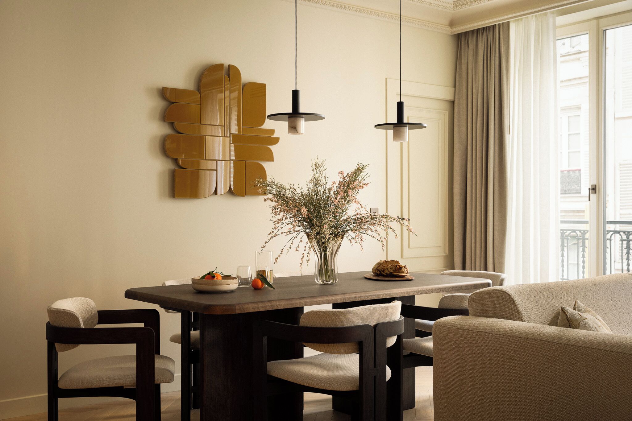 A modern dining area with a dark wooden table, two pendant lights, and a vase of flowers. There are chairs around the table and a sofa in the background.