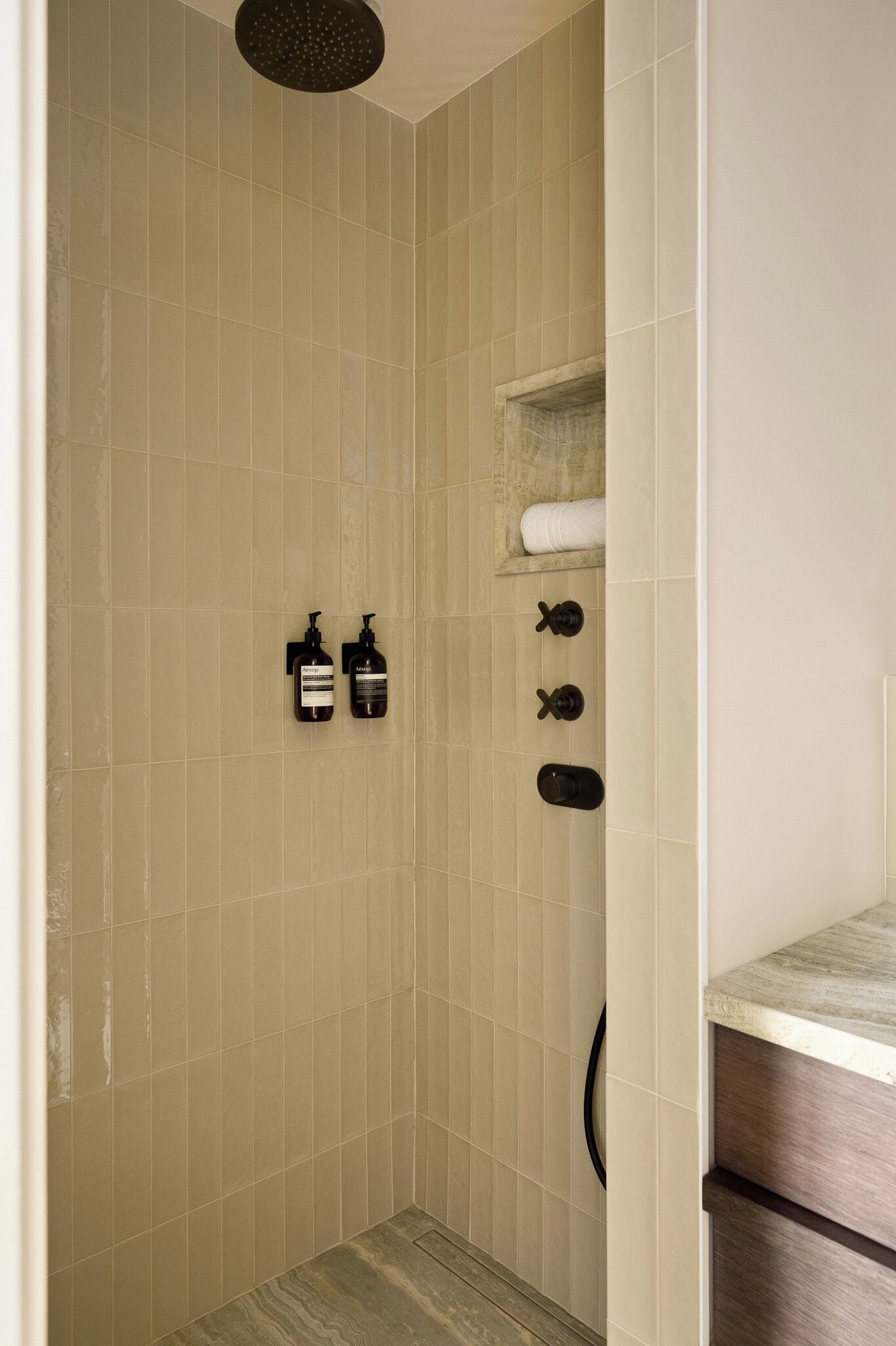 Une douche avec des carreaux de couleur beige, un pommeau de douche à main, un distributeur de savon et un porte-serviettes.