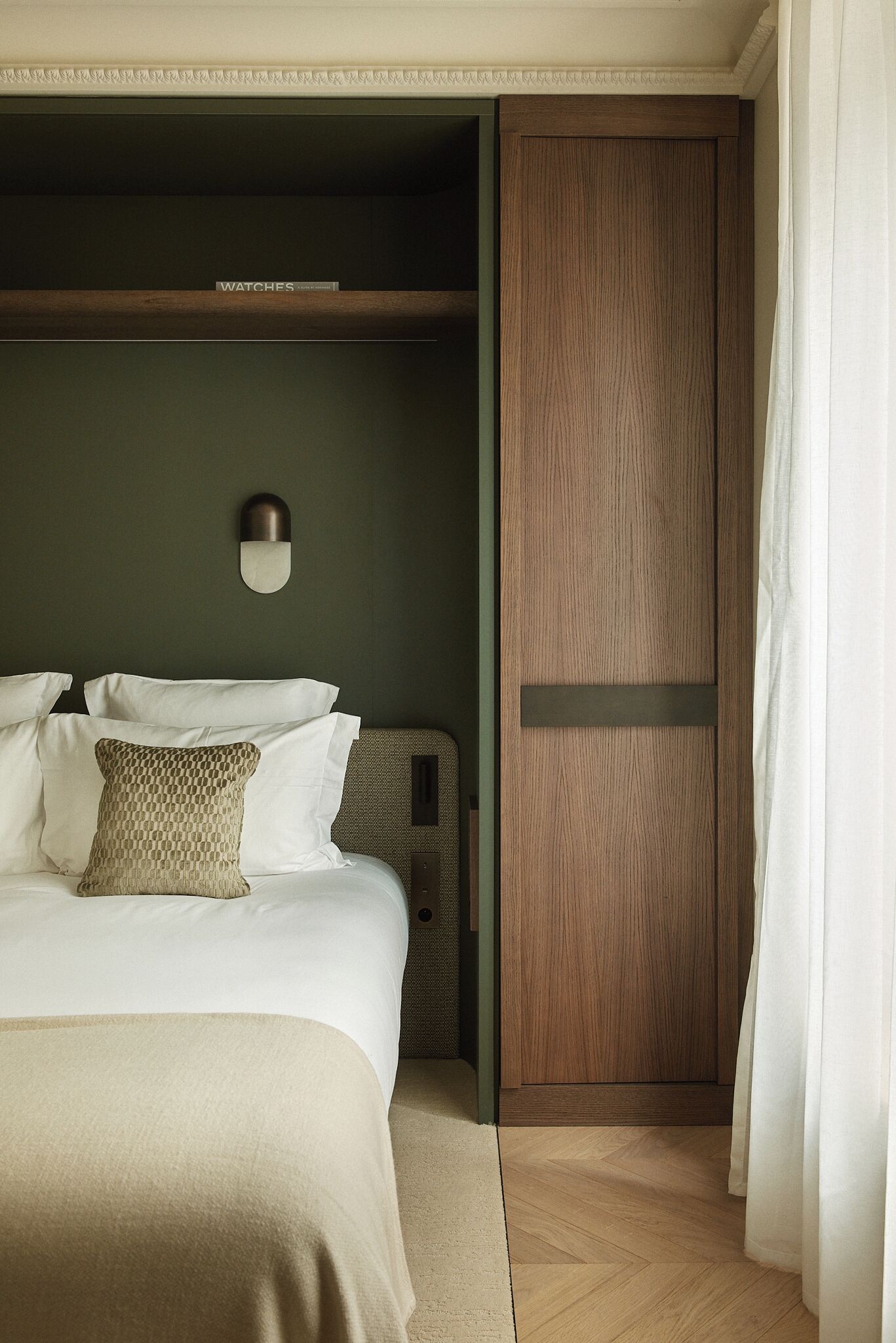A hotel room with a bed, a wooden wardrobe, and a wall-mounted light fixture.