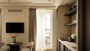 A hotel room with a flat-screen TV mounted on the wall, a small round table with a vase of flowers, two upholstered chairs, a window with sheer curtains, and built-in shelving with decorative items. - HIGHSTAY Maison - Boétie (Paris)