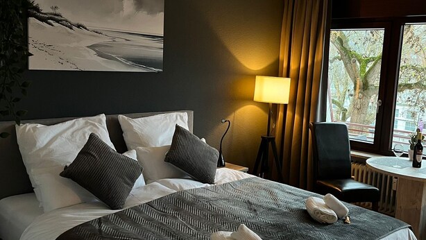 Un letto rifatto con cura, una coperta a motivi decorativi, un comodino con una lampada e un quadro appeso al muro.