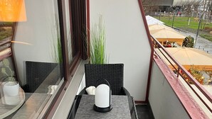 Un balcone con mobili in vimini, un tavolo e sedie. Si vede una finestra con vista su un edificio e degli alberi.
