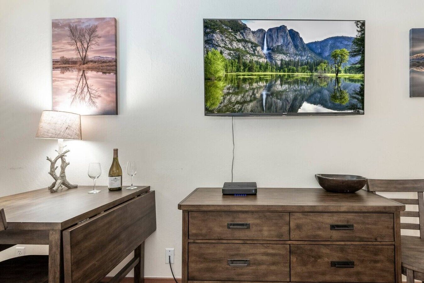 A dining area with a wooden table, a lamp, a wine bottle, and a flat-screen TV mounted on the wall.