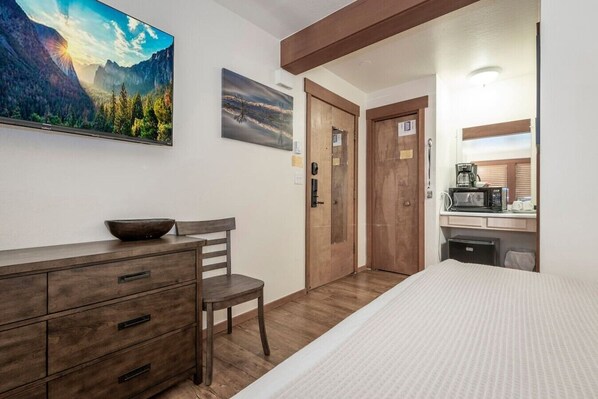 A bedroom with a bed, a wooden dresser, a chair, a desk with a microwave, and a view of a mountain landscape.