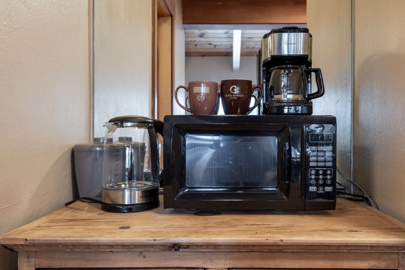Una kitchenette con cafetera, microondas y una jarra de vidrio sobre una encimera de madera.