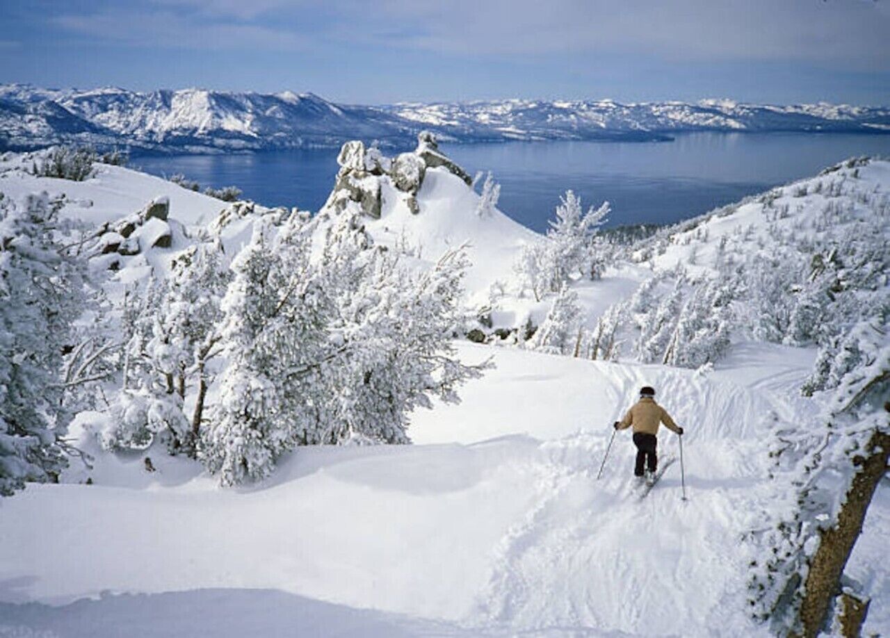 A snowy landscape with a person skiing, trees, and mountains in the background.