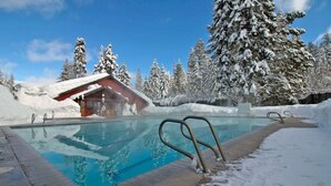 Una piscina con cubierta de madera, rodeada de árboles cubiertos de nieve y un edificio con techo inclinado.