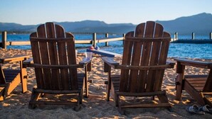 Two wooden Adirondack chairs on a sandy beach with a wooden table and a view of the ocean and mountains in the background.