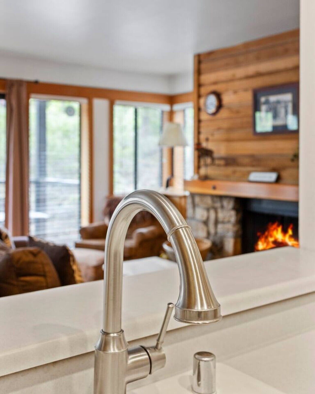 A modern kitchen with a stainless steel faucet, a white countertop, and a fireplace in the background.