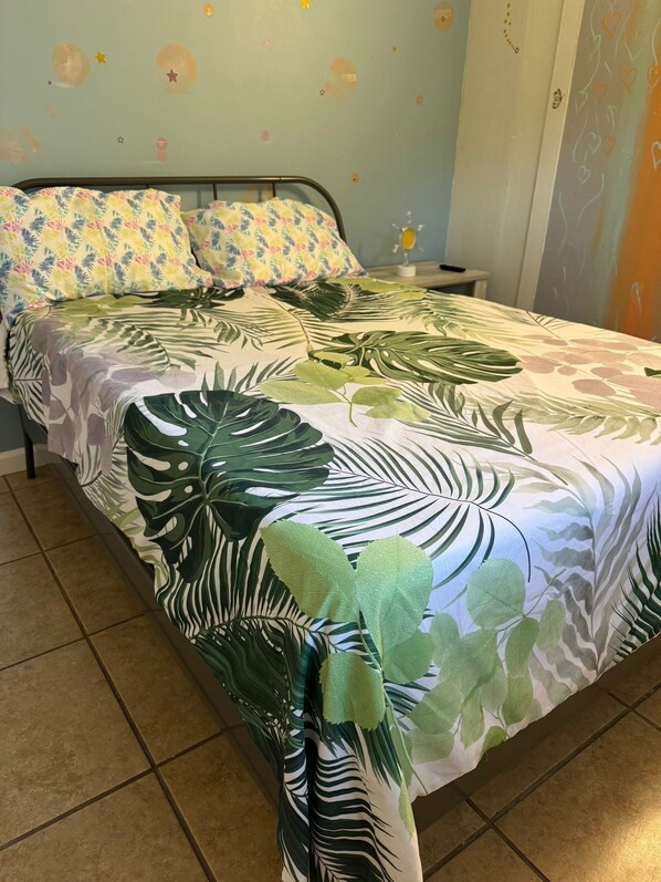A bed with a tropical leaf-patterned bedspread, a nightstand with a lamp, and a wall with star and heart decorations.