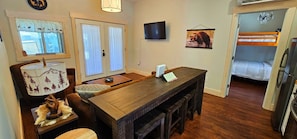 A compact kitchen with a wooden table, a TV mounted on the wall, and a bear-themed wall hanging.