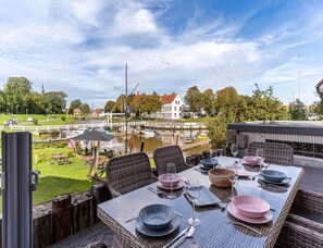Ein Essbereich im Freien mit Tischen für zwei Personen, mit Blick auf einen Kanal mit Booten und einer Brücke.