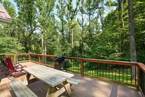 A wooden deck with a picnic table and chairs, surrounded by a metal railing, overlooking a forested area.