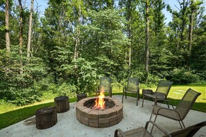 A circular fire pit with a fire burning, surrounded by outdoor furniture and a grassy area with trees in the background.