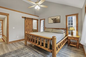 Ein Schlafzimmer mit einem hölzernen Bett, einem Deckenventilator, einem Nachttisch, einer Lampe und einem Fenster mit Vorhängen.