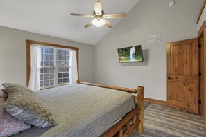 Ein Schlafzimmer mit Bett, Deckenventilator, Fenster, Fernseher und Holztür.