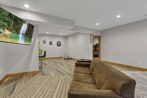A spacious living room with a large sectional sofa, a flat-screen TV mounted on the wall, and a hardwood floor.